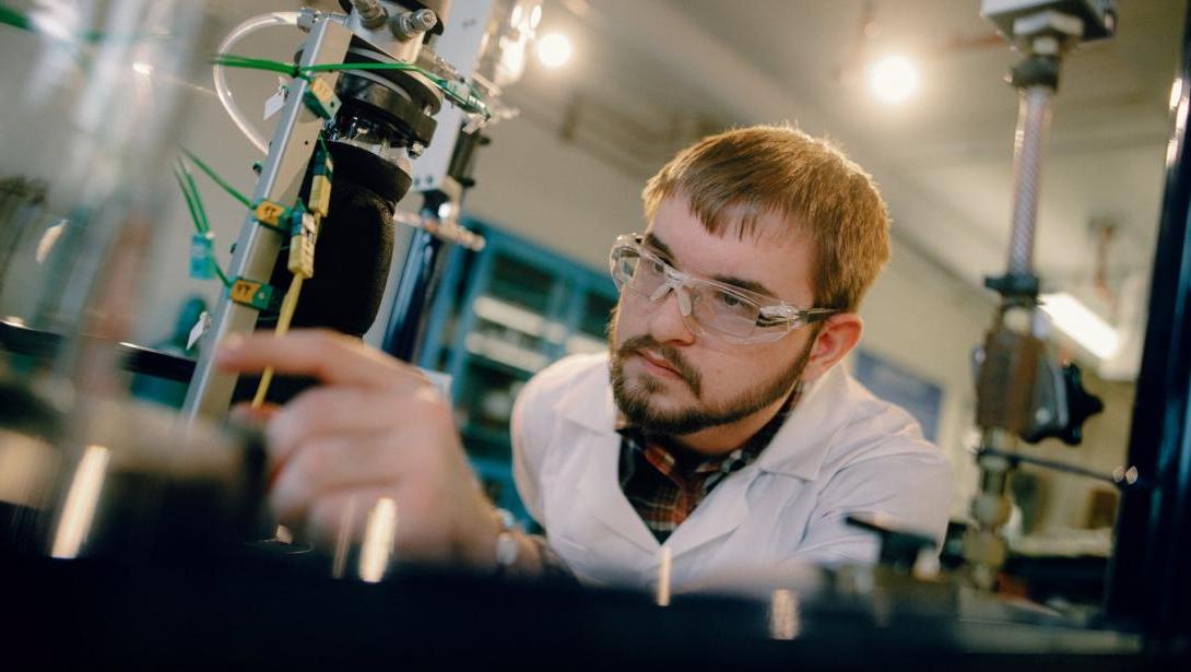 A student in a lab coat and goggles works with equipment