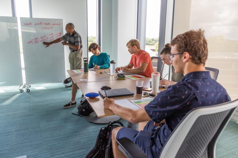 A faculty member guides students in an interactive study session using whiteboards in a dedicated meeting room within the Learning Commons.